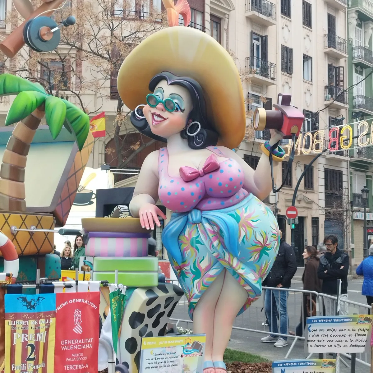 Ninot of a smiling tourist with a sun hat and camera beside prize banners at a Valencia falla